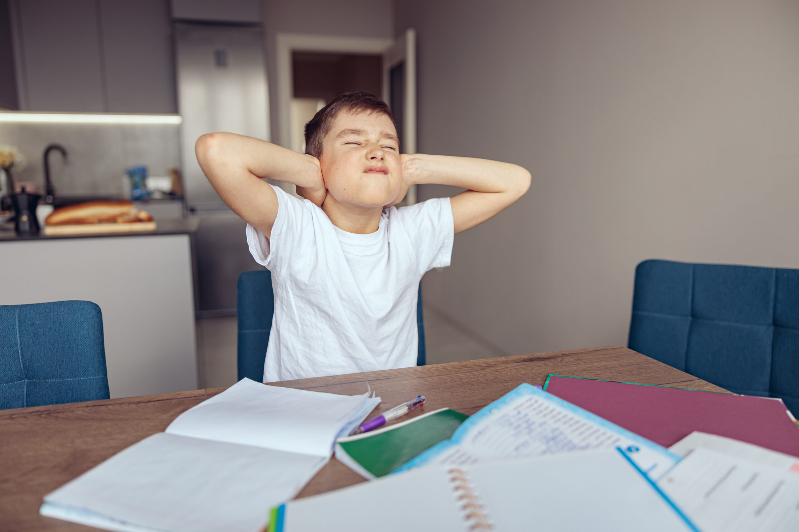 Caucasian small schoolboy closing ears with hands at homework and textbooks.
